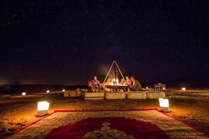 Desert Campfire Gathering Under Starry Sky Group sitting around a campfire at night in a desert with cushions, lanterns, and a patterned rug.