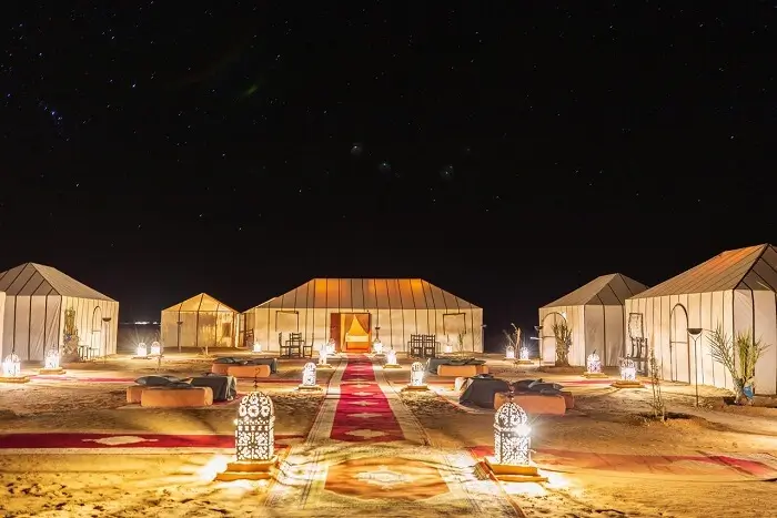 Desert camp with white tents and lantern-lit pathway at night Nighttime desert camp with illuminated white tents, lanterns, rugs, and low seating under a starry sky