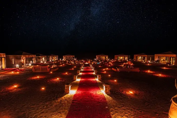 Lantern-lit Red Carpet Path to Tented Campsite at Night Nighttime desert camp with red carpet path, lanterns, fire pits, and tents under starry sky