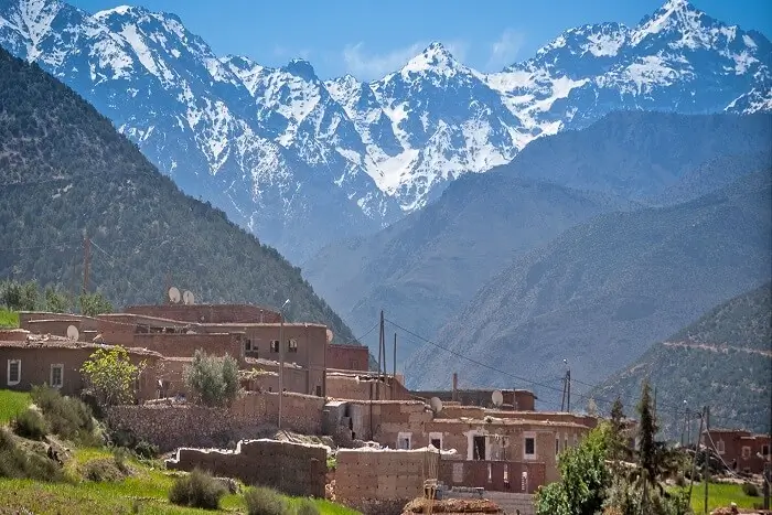 Village nestled between forested hills and snow-capped mountains Village with traditional buildings at forested hill base and snow-capped mountains behind.