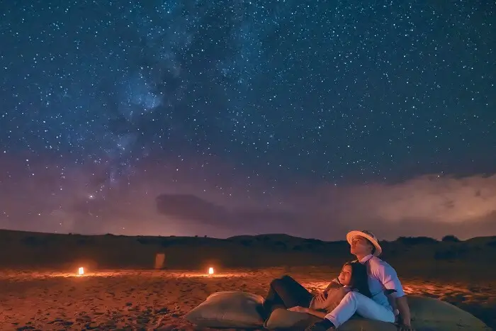 Couple Relaxing on Cushions Under Starry Night Sky Couple relaxing on cushions in sandy area at night with lanterns and starry sky above.