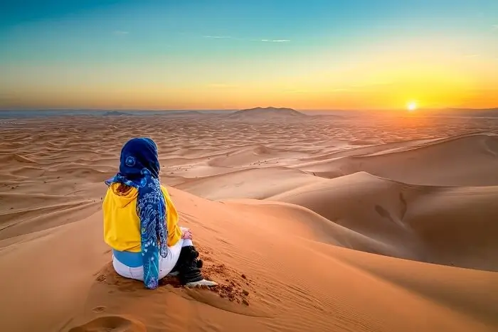 Person in yellow jacket on sand dune at sunset Person in yellow jacket and blue headscarf sitting on sand dune overlooking desert at sunset