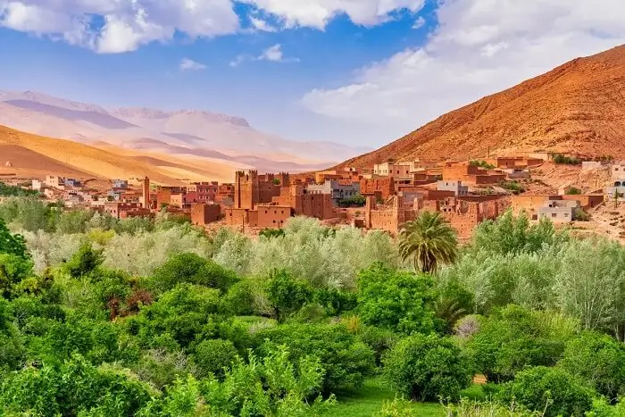 Village at the base of arid reddish mountains with green foreground vegetation Village with earthen buildings at the base of red mountains, lush greenery in foreground.