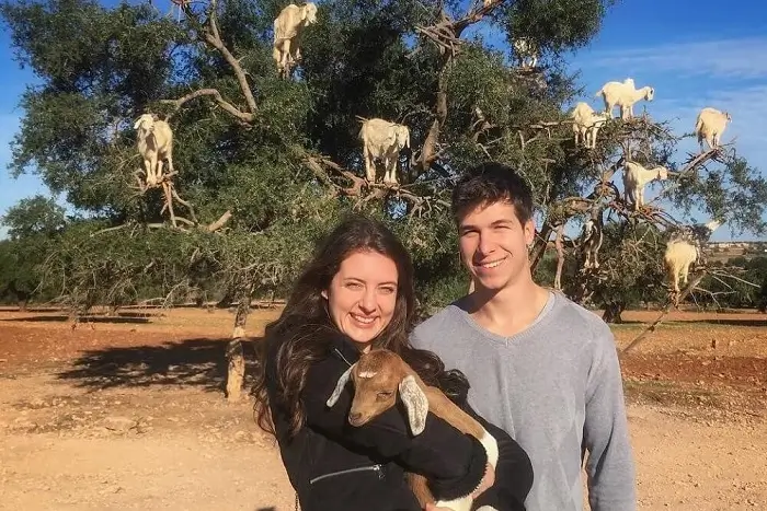 Smiling couple holding a baby goat in front of Argan tree climbing goats near Essaouira on their Morocco honeymoon